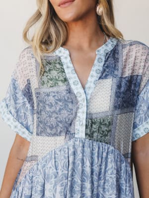 A close-up of a patchwork-print dress in soft blues, sage green, and blush — the kind of piece that looks like it was made from a collection of pretty vintage scarves, styled with gold shell earrings and a delicate chain necklace.