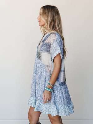 A flowy patchwork mini dress in soft blue tones worn in profile, layered with stacked turquoise bracelets — the kind of dress that looks equally at home on a cobblestone street or a sunny patio.
