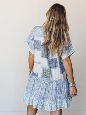 A back-facing model shows off a breezy patchwork mini dress in soft blue and white, its tiered ruffle hem and mixed floral and paisley print blocks giving it that effortlessly collected, vintage-quilt-meets-summer-dress energy.