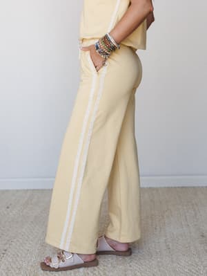 A side-angle shot of wide-leg pants in a soft butter yellow, detailed with a delicate lace side stripe — styled with a matching top, beaded bracelet stack, and strappy sandals for an easy, put-together feel.