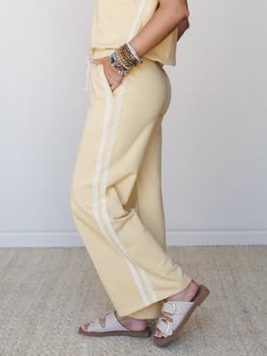 A side-profile shot of soft yellow wide-leg pants with a delicate lace side stripe, styled with matching yellow top, layered bead bracelets, and buckle sandals — the kind of easy, pulled-together set you reach for when comfort and cute need to coexis