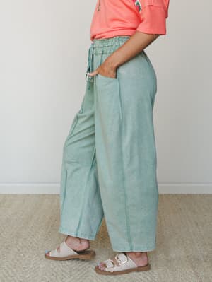 A side-profile shot of wide-leg jade green pants with a smocked waistband and relaxed silhouette, styled with a coral tee and neutral sandals for an easy, warm-weather feel.