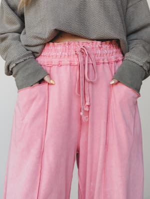 A close-up of wide-leg sweatpants in a washed dusty pink, styled with a cropped grey textured sweatshirt — the smocked waistband and drawstring tie give them a relaxed-but-intentional feel that's a step above your average cozy day.