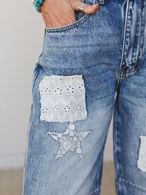 A close-up detail shot of medium-wash barrel jeans featuring a white eyelet patch and a lace star appliqué on the thigh, styled with turquoise beaded bracelets and a silver ring.