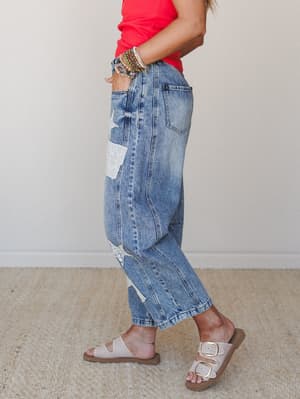 A side-profile shot of relaxed barrel jeans in medium-wash denim with eyelet and star-shaped patchwork details, styled with a coral tank and buckle sandals — the kind of effortless outfit that looks like it came together without trying.
