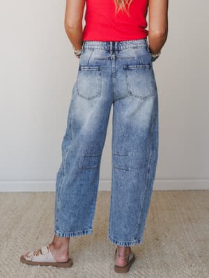 Back view of a model in wide-leg barrel jeans with a light acid wash and visible patch seaming at the knee, paired with a red ribbed tank and cream buckle sandals — relaxed and lived-in in the best way.