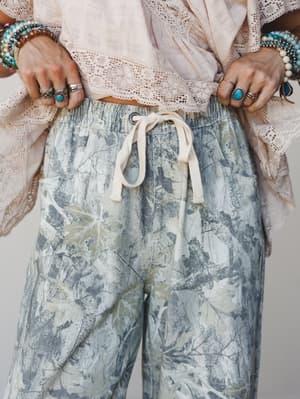 A close-up torso shot styled around relaxed tie-dye pants with a botanical, watercolor-like print — paired with a lace-trim crop top and stacked turquoise and silver jewelry that leans deep into boho territory.