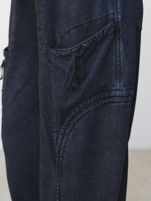 A close-up detail shot of deep navy-black cargo-style pants, highlighting a utilitarian side pocket with a drawstring tie and brass grommet hardware — worn-in texture gives them that lived-in, effortlessly cool feel.