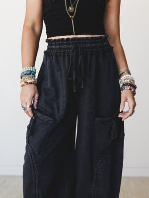A close-up torso shot of wide-leg black cargo pants paired with a smocked black crop top, layered gold necklaces, and both wrists stacked with colorful beaded bracelets — relaxed and expressive with an effortlessly boho edge.