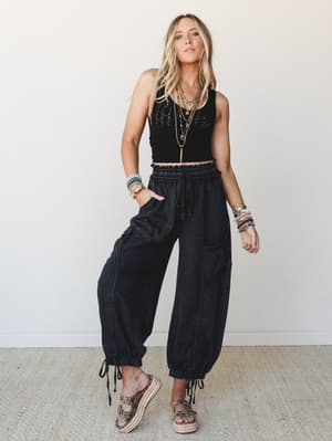 A full-length model shot of an all-black outfit — smocked crop tank paired with wide-leg cargo joggers — styled with layered gold necklaces, stacked bracelets, and platform espadrille sandals against a clean white studio wall.