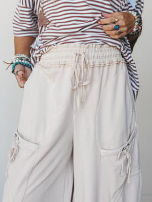 A close-up torso shot focused on wide-leg cargo-style pants in a soft oat cream, paired with a striped tee and layered beaded bracelets — relaxed and lived-in with just enough detail to make it interesting.