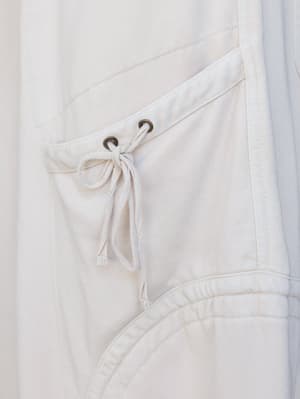 A close-up detail shot of soft oat-colored pants, highlighting a drawstring tie with brass grommets, a patch pocket, and gathered elastic detailing — relaxed and intentional all at once.
