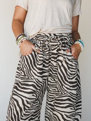A cropped torso shot of wide-leg zebra print pants styled with a knotted cream tee and stacked beaded bracelets — effortless and a little wild in the best way.