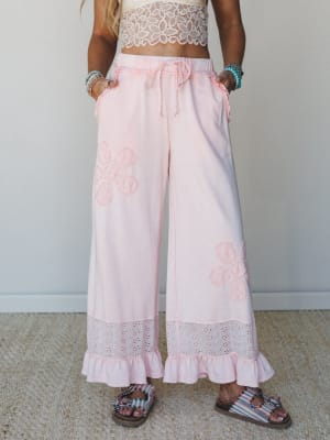 Dreamy ice pink wide-leg pants with crochet flower appliqués, eyelet hem trim, and a ruffle cuff — styled with a lace crop top and stacked bracelets for a look that feels like summer decided to stay.