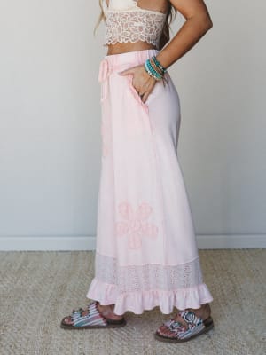 A side-profile shot of a soft pink maxi skirt with tonal floral appliqué and an eyelet ruffle hem, paired with a cream lace crop top and printed sandals — the whole look reads like a summer afternoon that never quite ends.