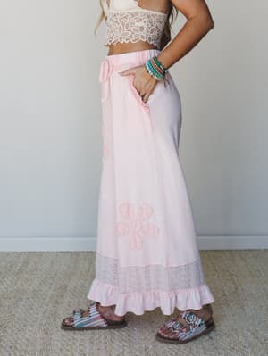 A side-profile shot of a soft pink maxi skirt with tonal floral appliqué and an eyelet ruffle hem, paired with a cream lace crop top and printed sandals — the whole look reads like a summer afternoon that never quite ends.