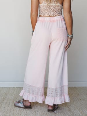 A back-view shot of wide-leg soft pink pants with eyelet hem trim and ruffle detail, paired with a cream crochet crop top and printed sandals — relaxed and feminine with a boho-sweet energy.