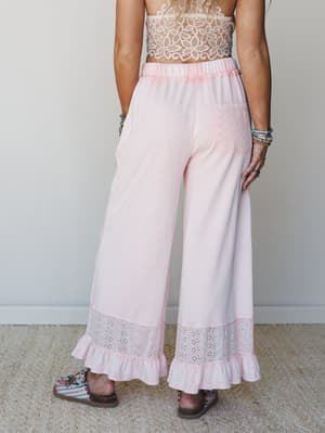A back-view shot of wide-leg soft pink pants with eyelet hem trim and ruffle detail, paired with a cream crochet crop top and printed sandals — relaxed and feminine with a boho-sweet energy.