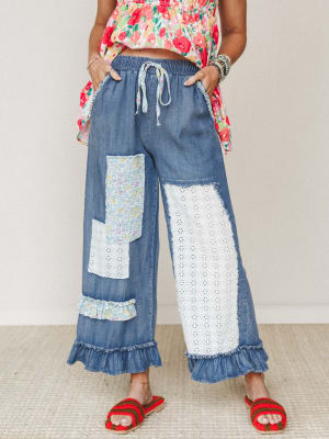 Wide-leg denim pants with patchwork eyelet and floral fabric panels, ruffle hem, and a fabric drawstring waist — styled with a bright floral top and crochet sandals for a playful, lived-in boho look.