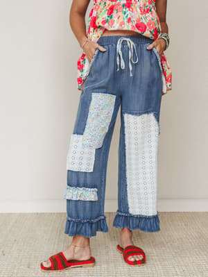 Patchwork denim wide-leg pants with eyelet panels, floral fabric patches, and ruffle hem details — styled with a bright floral top and red crochet sandals for a playful, lived-in boho look.