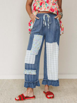Patchwork denim wide-leg pants with eyelet panels, floral fabric patches, and ruffle hem details — styled with a bright floral top and red crochet sandals for a playful, lived-in boho look.