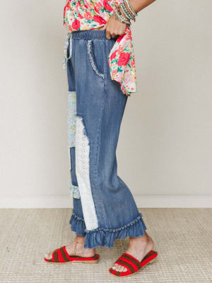 A side-profile shot of wide-leg denim pants with patchwork panels, eyelet lace trim, and a ruffled hem — paired with a bright floral top and red crochet sandals for a look that feels like a Sunday market and a good mood rolled into one.