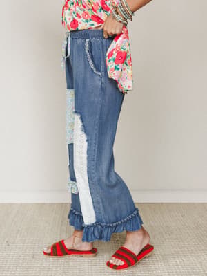 A side-profile shot of wide-leg denim pants with patchwork panels, eyelet lace trim, and a ruffled hem — paired with a bright floral top and red crochet sandals for a look that feels like a Sunday market and a good mood rolled into one.