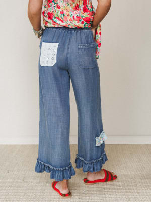 A back-view shot of wide-leg chambray pants with ruffled hems, an eyelet lace patch pocket, and a small floral fabric bow at the ankle — styled with a red floral smocked top and red woven sandals for a playful, garden-ready look.