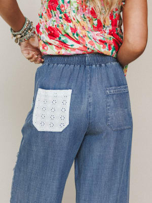 A close-up back view of relaxed wide-leg denim pants with a standout eyelet patch pocket, styled with a floral smocked top and stacked boho bracelets — the kind of outfit that feels effortless but clearly thought about.
