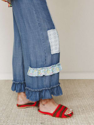 A close-up of wide-leg dark denim pants with layered ruffle hems, an eyelet patch detail, and a floral ruffle inset — paired with red and brown crochet slide sandals for a playful, handmade-feeling finish.