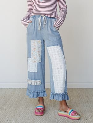 Wide-leg light denim pants with eyelet lace and floral fabric patches, ruffled hems, and a fabric drawstring waist — styled with a pink striped long-sleeve and rainbow platform sandals for a playful, lived-in boho feel.
