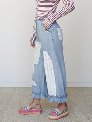 A side-profile shot of wide-leg light denim pants with playful patchwork, eyelet lace panels, and a ruffled hem, styled with a pink striped long-sleeve and rainbow platform sandals against a soft neutral interior.