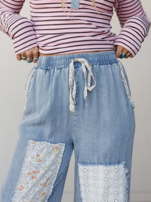 A close-up of easy wide-leg denim pants styled with a pink striped long-sleeve top — the pants are the real story, with patchwork floral and eyelet panels, a smocked elastic waistband, and a floral fabric drawstring that feel like they were stitched