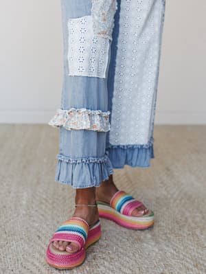 A close-up of wide-leg light denim pants with mixed eyelet and floral ruffle trim at the hem, paired with rainbow stripe crochet platform sandals on a jute rug — the kind of outfit that makes even a Tuesday feel intentional.