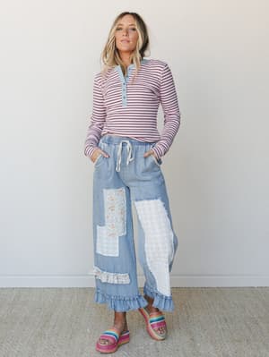 A wide-leg light denim pant with patchwork eyelet and floral details steals the show, styled with a pink striped henley and rainbow platform sandals against a clean white studio backdrop — casual and expressive all at once.