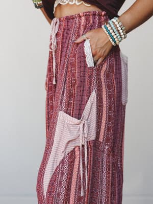 A close-up detail shot of wide-leg boho print pants in deep mauve and blush, styled with stacked beaded bracelets and a lace-trim crop top — the kind of pants that make a Tuesday feel intentional.
