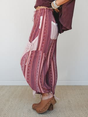 A side-profile shot of billowy harem-style pants in a warm mauve mixed print, styled with a cropped burgundy top, wood-platform clogs, and stacked turquoise bracelets — the kind of outfit that feels intentional and well-traveled at the same time.