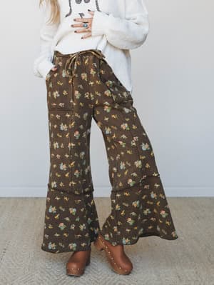 Wide-leg floral pants in a rich chocolate brown ditsy print, styled with a chunky cream sweater and tan clogs — the kind of outfit that feels like fall finally got interesting.