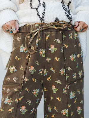 A close-up waist-down shot of brown floral cargo-style pants with a self-tie drawstring, styled with a cream chunky knit sweater and layered gingham necklace — earthy, vintage, and very much the pants you'd be emotionally attached to.