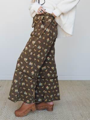 Wide-leg floral pants in a deep brown with scattered ditsy bouquets are styled with a chunky cream sweater and studded wood clogs — the kind of outfit that feels vintage-inspired without trying too hard.