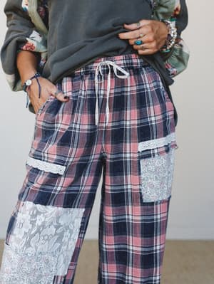 A close-up mid-shot of wide-leg plaid pants in dusty rose and navy, detailed with mixed lace and floral patchwork panels — styled casually with a faded charcoal sweatshirt and layered boho jewelry for a relaxed, collected look.