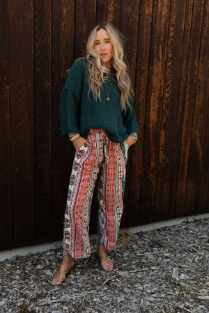 A woman with long blonde hair wears The Nest Staycation Wide Leg Drawstring Pant in Red Clay, a dark green sweater, sandals, and layered jewelry, posing against a wooden wall with hands in her pockets for a relaxed look.
