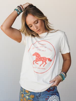 A relaxed ivory graphic tee with a faded red running horse print and 'Stay Wild, Keep Dreaming' text, styled with embroidered jeans and stacked beaded bracelets — the kind of outfit that feels effortless but says everything.