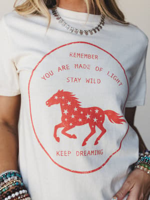 A cream graphic tee worn with stacked beaded bracelets and a silver coin necklace — the distressed red horse print reads 'Remember You Are Made of Light, Stay Wild, Keep Dreaming,' and the whole thing feels like a love letter to free-spirited dressin