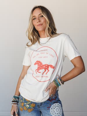 A sun-kissed model in an ivory vintage-graphic tee and heavily embroidered jeans, stacked with beaded bracelets — the kind of outfit that looks effortless but says everything about who she is.