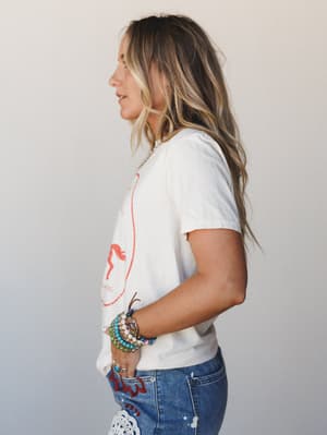 A side-profile shot of a model in an ivory graphic tee tucked loosely into embroidered patchwork denim, stacked with colorful beaded bracelets — the kind of effortless outfit that looks like it came together without trying.