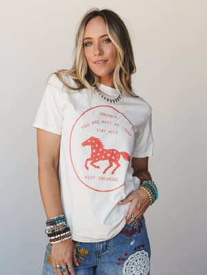 A relaxed ivory graphic tee with a distressed red horse-and-stars motif styled with embroidered denim and stacked boho jewelry — the kind of outfit that feels equally at home at a farmers market or a country concert.