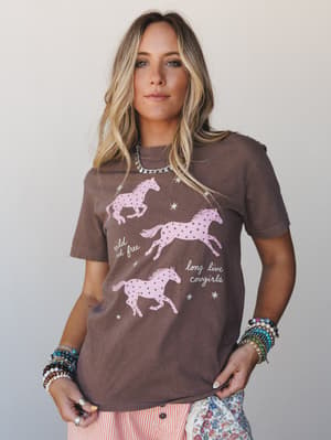A relaxed graphic tee in warm espresso brown featuring three pink galloping horses with star details and the text 'wild and free' and 'long live cowgirls', styled with stacked beaded bracelets, a charm necklace, and a peek of floral and striped botto