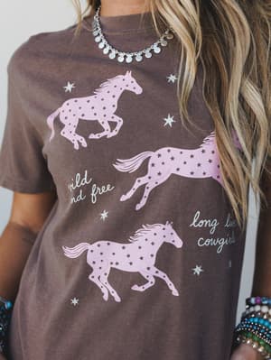 A close-up of an espresso brown graphic tee featuring three pink galloping horses with star details and the phrases 'wild and free' and 'long live cowgirls,' styled with a silver coin necklace and colorful bead bracelets.