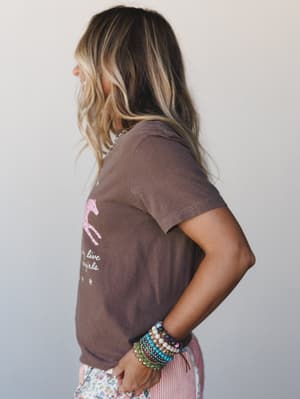A side-profile shot of a model in an espresso brown graphic tee featuring a pink horse illustration and 'long live cowgirls' text, styled with stacked beaded bracelets and a peek of floral and striped bottoms — effortlessly casual with a cowgirl-boho